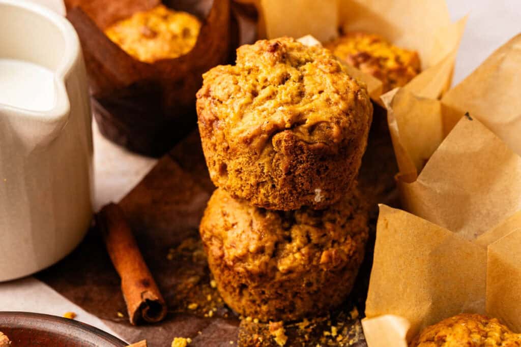 Two Vegan Pumpkin Muffins are stacked on a wooden board, surrounded by more muffins in parchment wrappers, with a cinnamon stick and a jug of milk nearby.