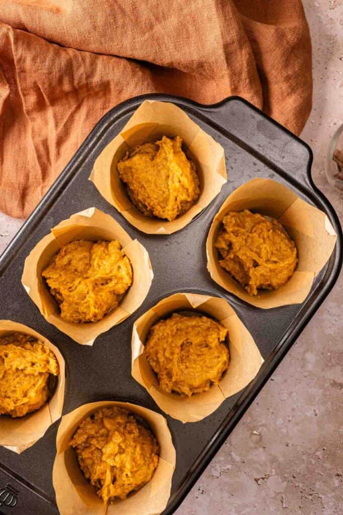 A muffin tin filled with six sections of unbaked Vegan Pumpkin Muffins batter in parchment paper liners, placed on a light countertop with a brown cloth nearby.