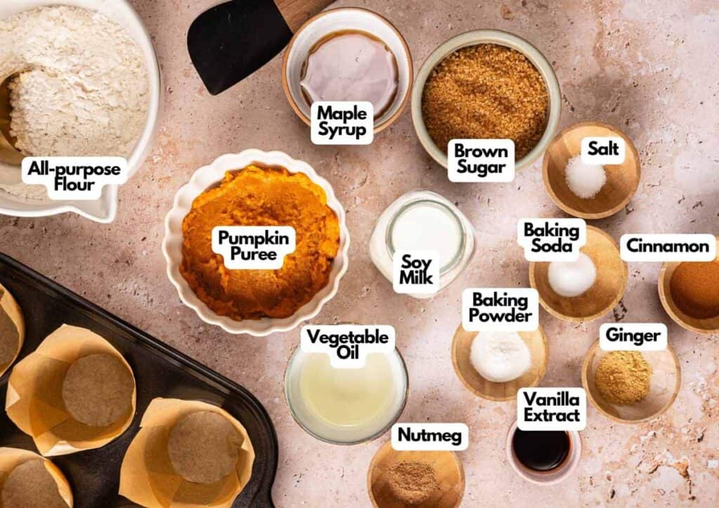 Top-down view of labeled baking ingredients for Vegan Pumpkin Muffins in bowls on a countertop, including flour, pumpkin puree, maple syrup, plant milk, oil, sugar, spices, salt, and leavening agents.