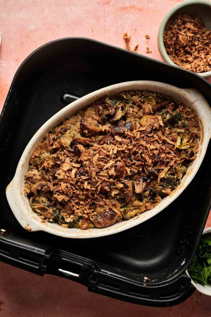 An Air Fryer Vegan Broccoli Casserole topped with crispy fried onions and mushrooms sits in an oval dish inside a black air fryer basket.