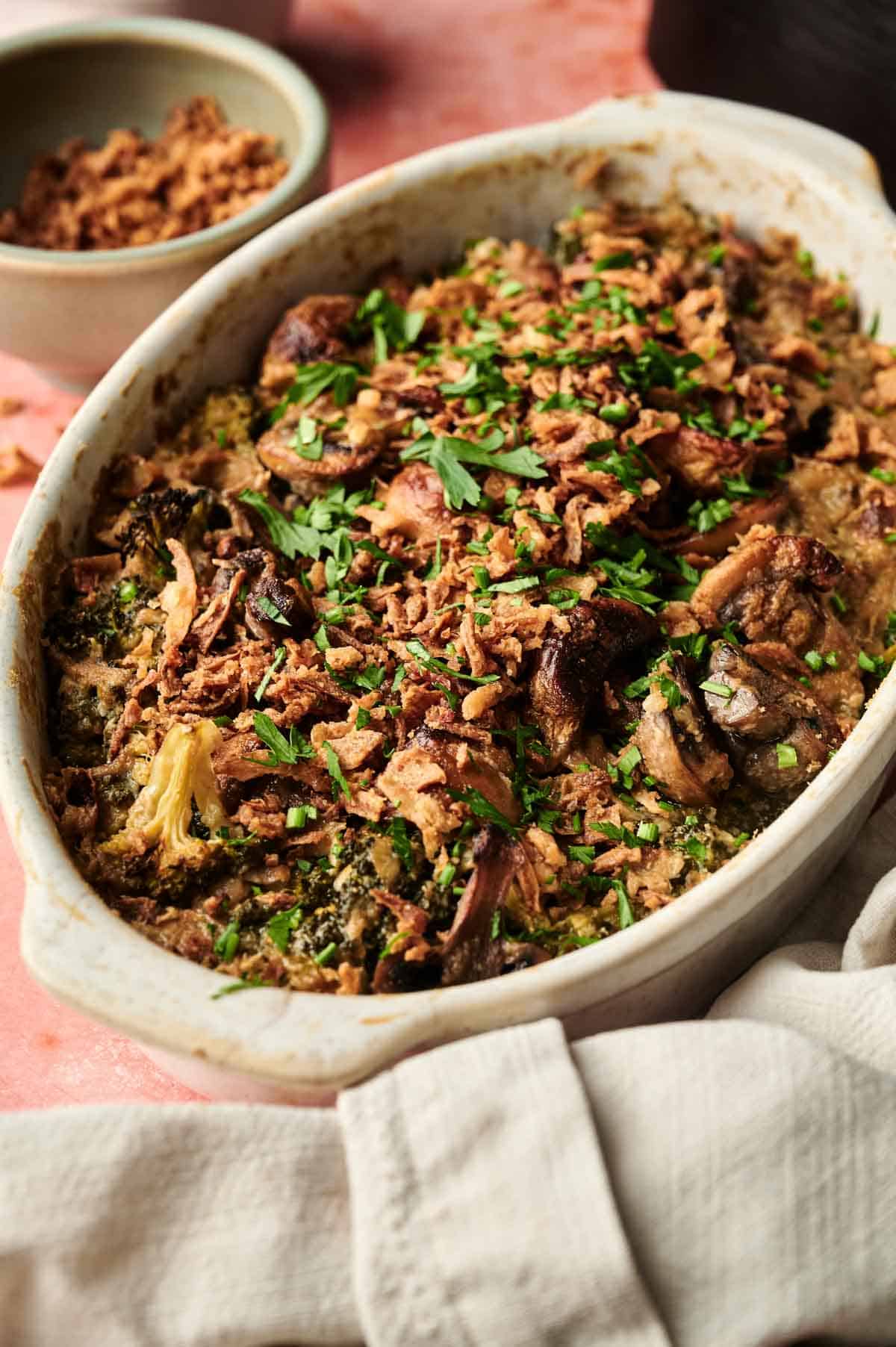 An oval baking dish filled with an Air Fryer Vegan Broccoli Casserole, topped with crispy onions and chopped fresh herbs, set on a pink surface with a napkin nearby.