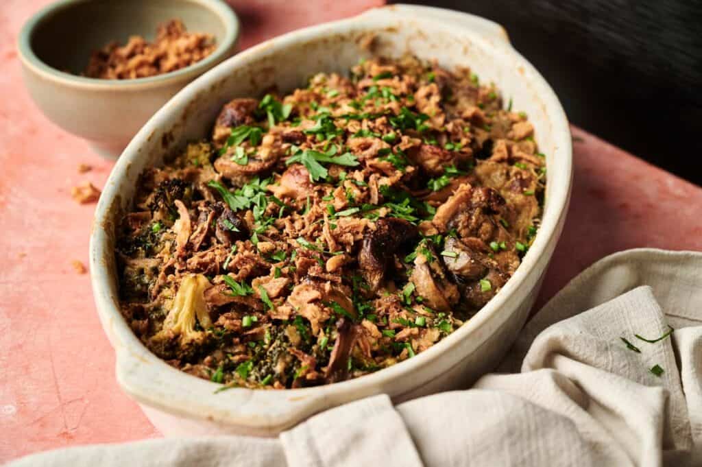 An Air Fryer Vegan Broccoli Casserole topped with chopped herbs and crispy fried onions sits on a pink surface next to a bowl of extra onions and a folded cloth napkin.