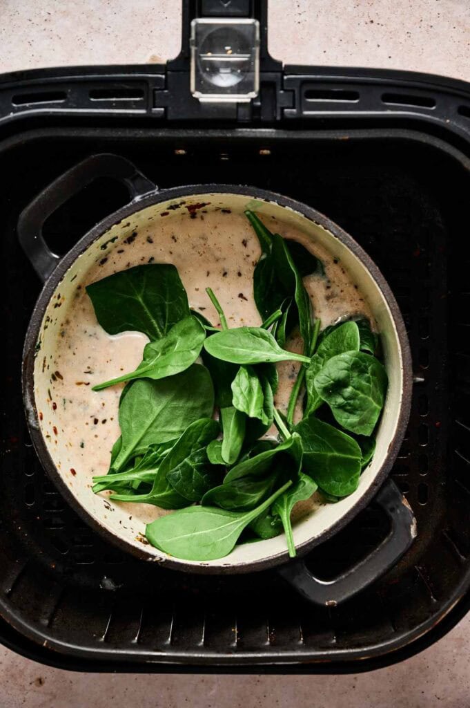 A pot with creamy sauce and fresh spinach leaves sits inside an air fryer basket, the perfect start for Air Fryer Marry Me Chickpeas on a light countertop.