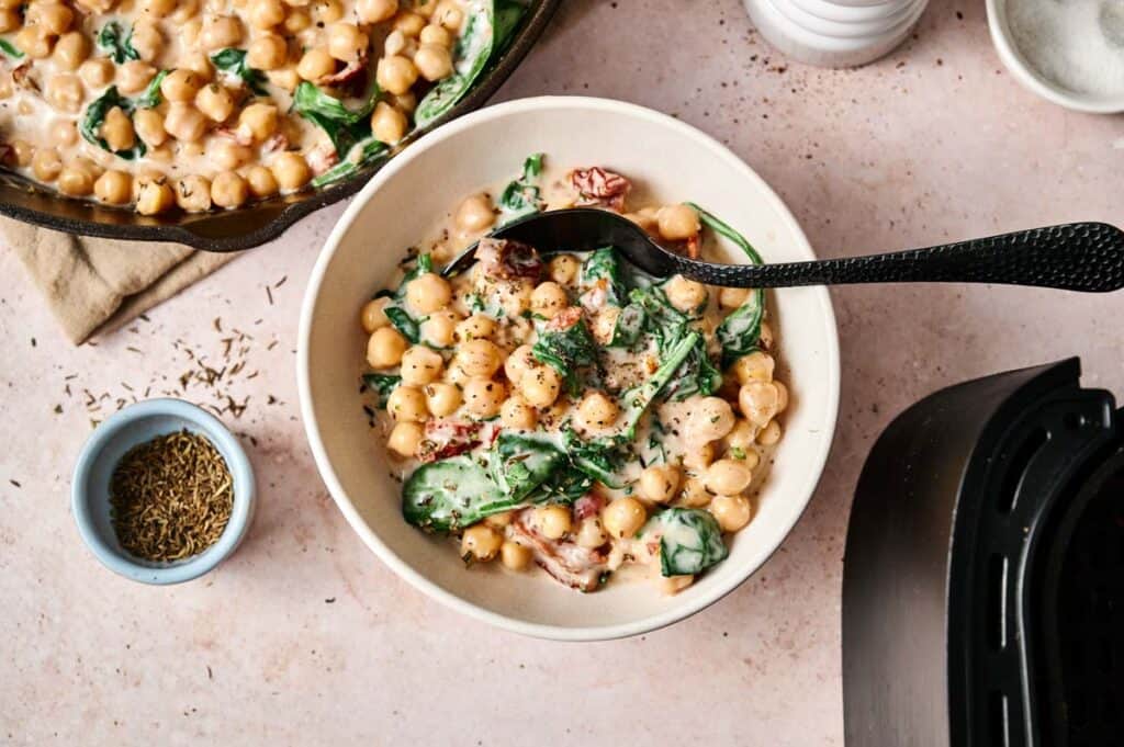 A bowl of creamy chickpea and spinach stew with herbs, inspired by Air Fryer Marry Me Chickpeas, is garnished with parsley and sits on a pink surface beside a skillet, a small bowl of dried herbs, and a pepper grinder.