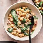 A bowl of creamy Air Fryer Marry Me Chickpeas and spinach stew with a black spoon, garnished with herbs. A small dish of dried herbs is nearby.