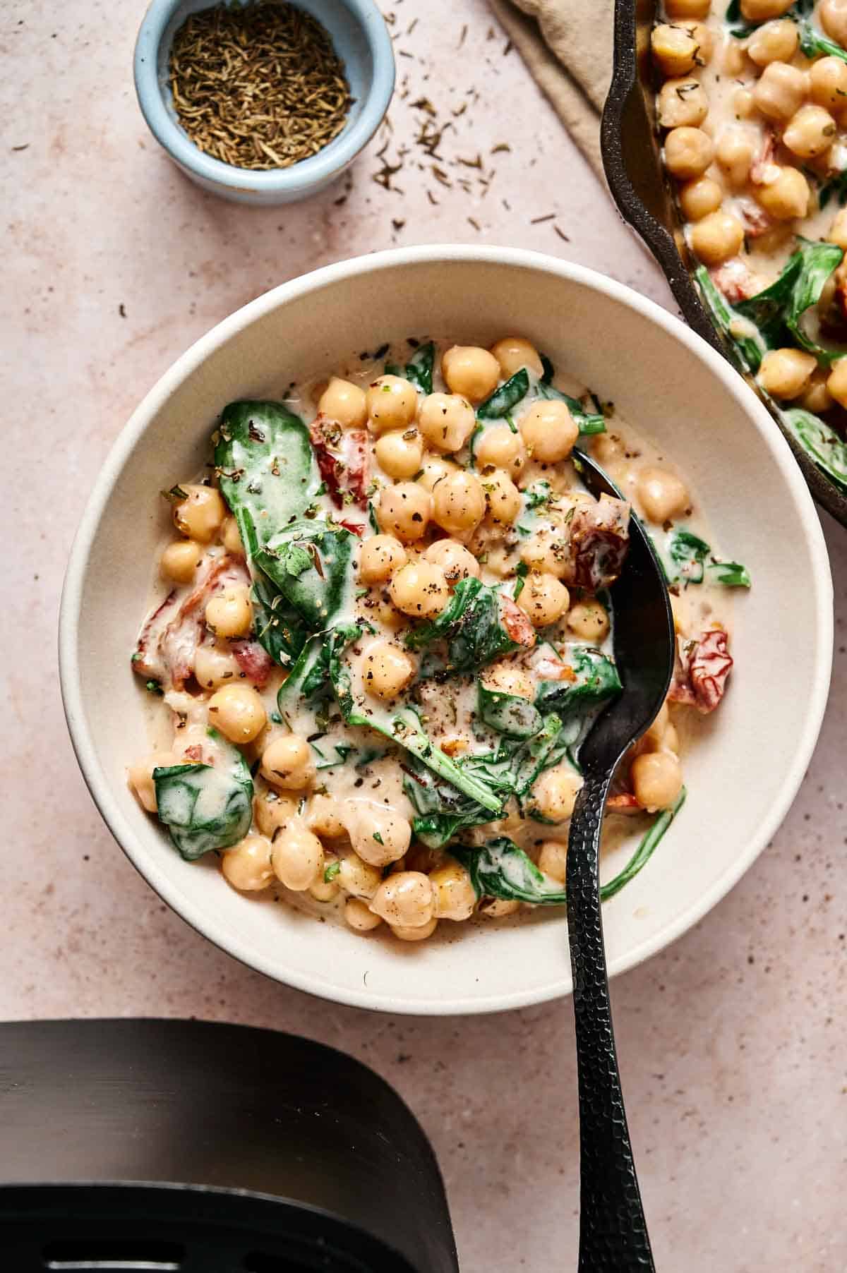 A bowl of creamy Air Fryer Marry Me Chickpeas and spinach stew with a black spoon, garnished with herbs. A small dish of dried herbs is nearby.