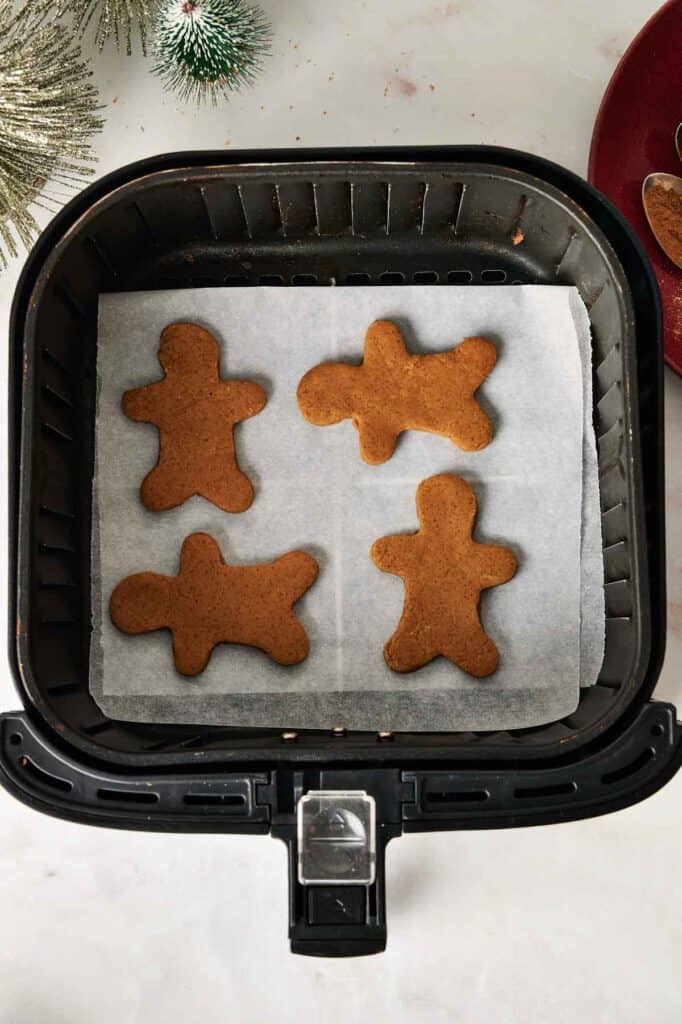 Four Air Fryer Vegan Gingerbread Cookies shaped like people are arranged on parchment paper inside an air fryer basket.