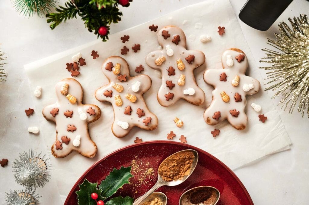 Three Air Fryer Vegan Gingerbread Cookies decorated with small bear and mitten sprinkles are displayed on parchment paper, surrounded by holiday decorations and spices.