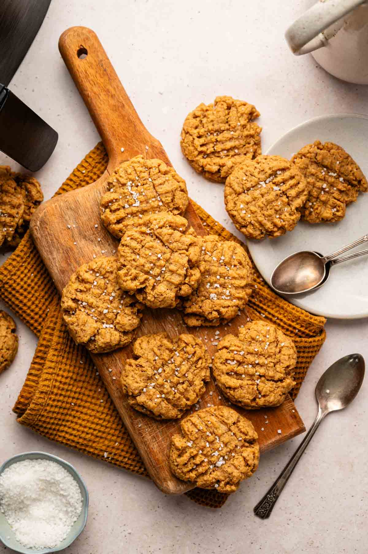 A wooden board with several Air Fryer Vegan Peanut Butter Cookies sprinkled with sea salt, placed on a brown cloth, surrounded by plates, spoons, and a small bowl of salt.