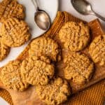 Air Fryer Vegan Peanut Butter Cookies with a crisscross pattern are arranged on a wooden board and plate, sprinkled with coarse salt. Spoons and a textured cloth are nearby.