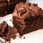 A slice of Vegan Chocolate Cake with thick chocolate frosting sits on a plate, with a fork holding a bite-sized piece in the foreground.