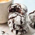 A stack of four Vegan Crinkle Cookies dusted with powdered sugar, with the top cookie partially bitten, sits next to a cup of coffee.