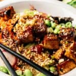 A Vegan Poke Bowl featuring tofu cubes, edamame, shredded carrots, radish slices, scallions, and sesame seeds, with black chopsticks resting on the side.