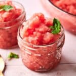 Two small glass jars filled with refreshing watermelon sorbet granita, each topped with fresh mint leaves, are placed on a light surface.