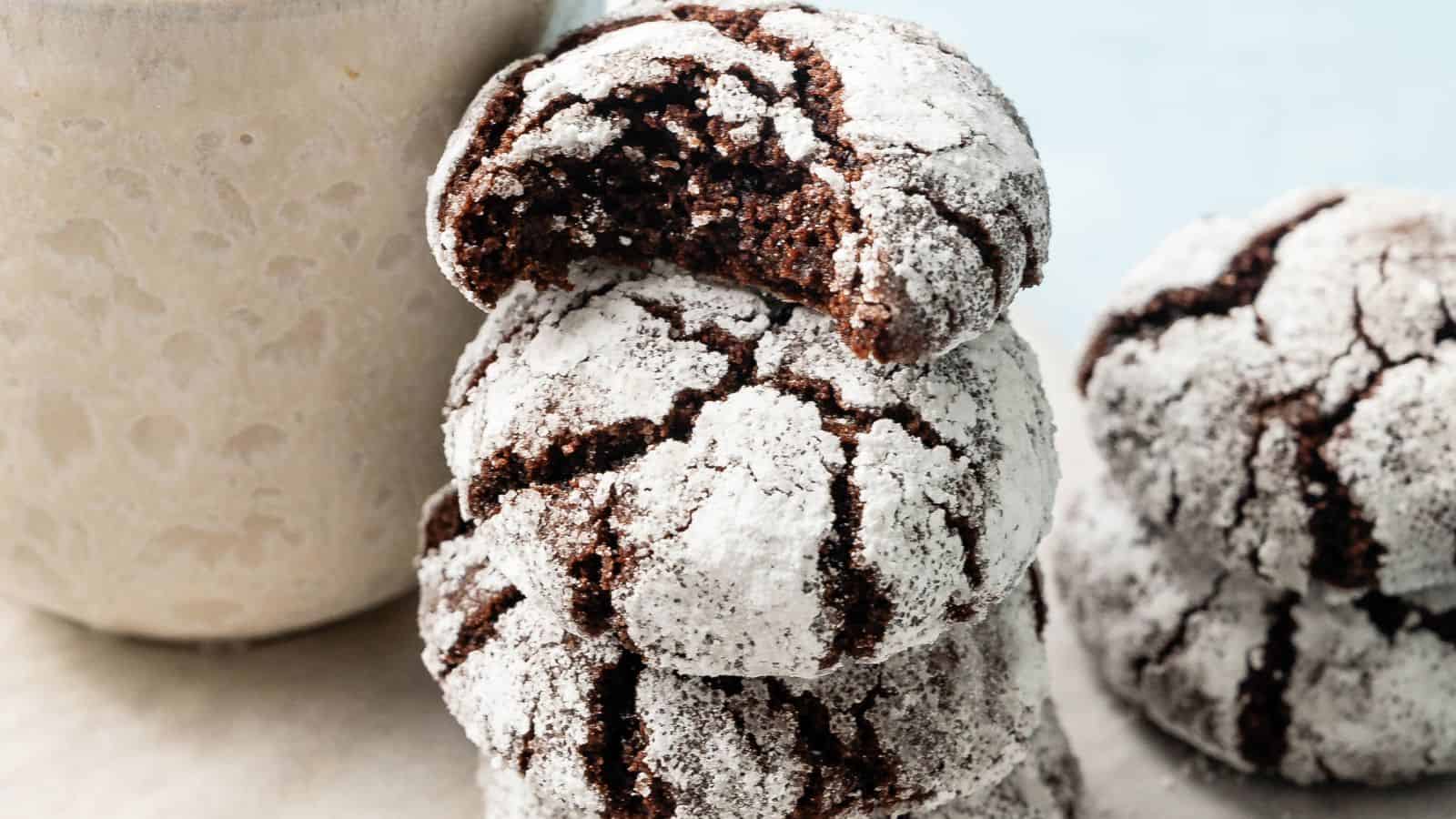 A stack of powdered sugar-coated chocolate crinkle cookies, with one cookie on top showing a bite taken out, next to a light-colored cup.