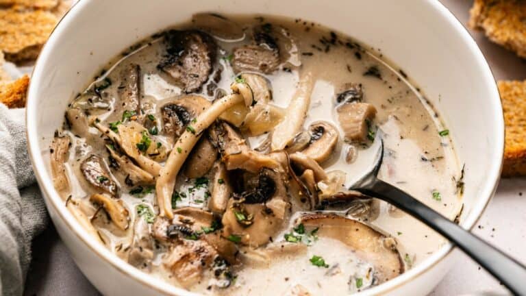 A bowl of creamy mushroom soup with sliced mushrooms, herbs, and a spoon, surrounded by pieces of bread.