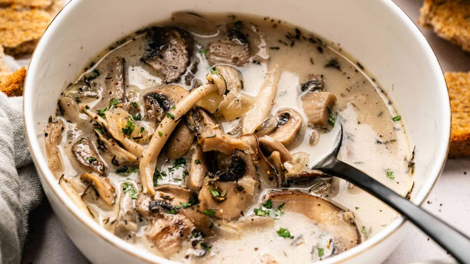 A bowl of creamy mushroom soup with sliced mushrooms, herbs, and a spoon, with pieces of bread on the side.