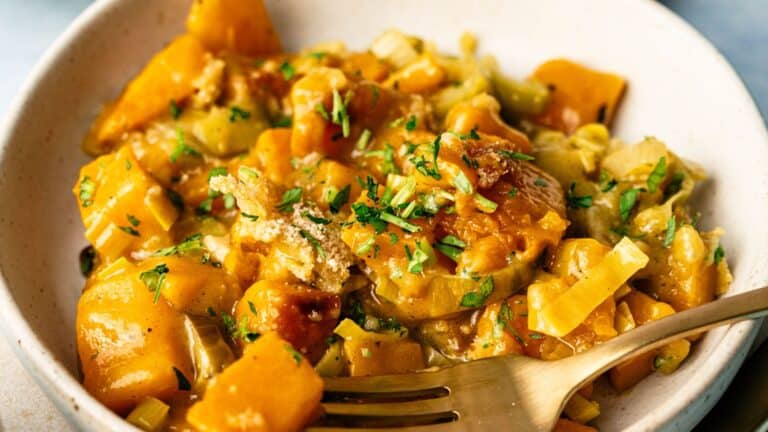 A bowl of cooked vegetables, primarily chunks of butternut squash, garnished with chopped herbs and served with a fork.