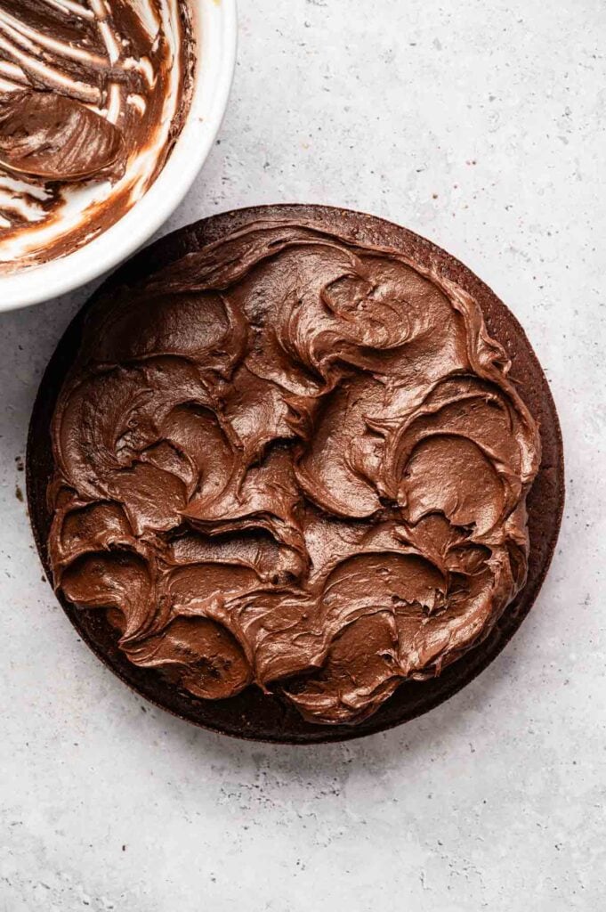 A round Vegan Chocolate Cake with swirled chocolate frosting on top sits next to a bowl with remnants of rich, creamy frosting.