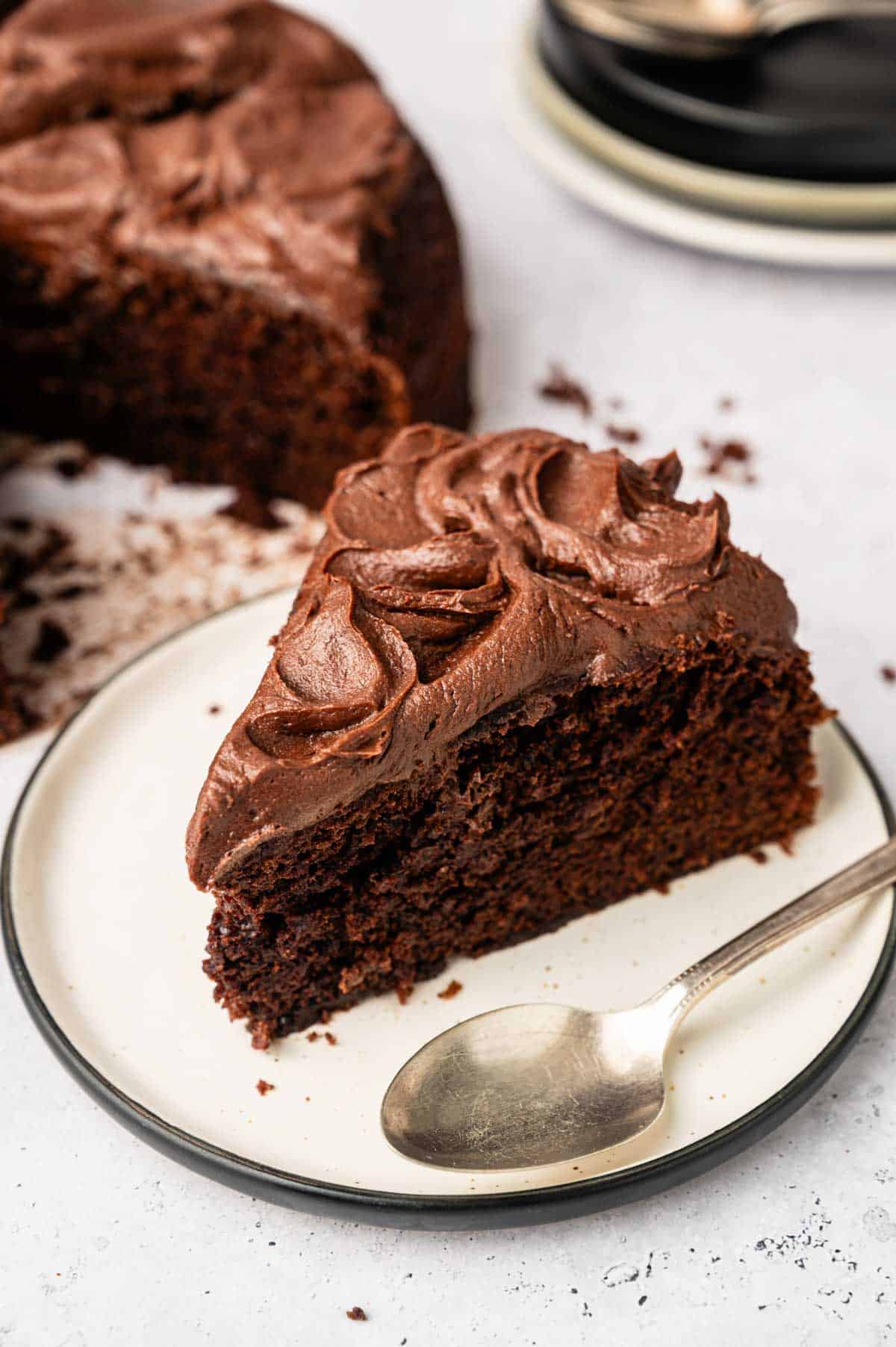 A slice of Vegan Chocolate Cake with chocolate frosting sits on a white plate next to a spoon, with the rest of the cake and stacked plates in the background.