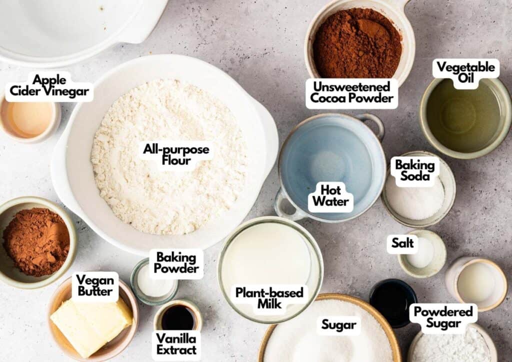 Bowls containing all-purpose flour, unsweetened cocoa powder, and other ingredients set the stage for a delicious Vegan Chocolate Cake made with plant-based milk, apple cider vinegar, vegan butter, and vanilla extract.