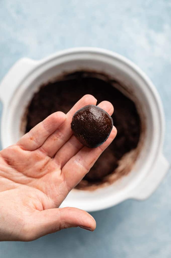 A hand holds a round ball of chocolate dough above a white bowl containing more dough, ready to become delicious Vegan Crinkle Cookies, all set on a light blue surface.