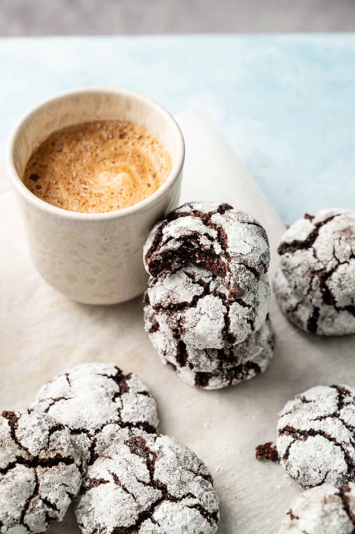 A cup of coffee sits next to a stack of Vegan Crinkle Cookies, dusted with powdered sugar, on a light surface.