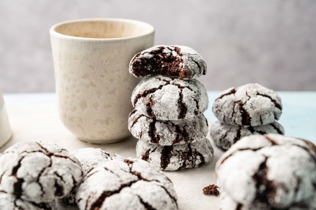 A stack of Vegan Crinkle Cookies coated in powdered sugar sits next to a beige ceramic cup on a light surface.