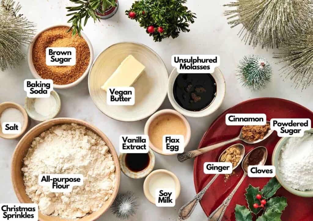 Assorted labeled baking ingredients for Air Fryer Vegan Gingerbread Cookies, including flours, sugars, spices, vegan butter, soy milk, flax egg, and decorative Christmas sprinkles, arranged on a festive surface.