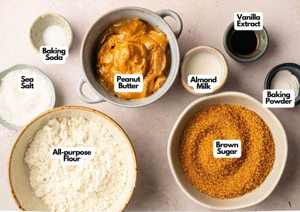 Bowls containing labeled ingredients for baking Air Fryer Vegan Peanut Butter Cookies&mdash;peanut butter, brown sugar, all-purpose flour, almond milk, vanilla extract, baking powder, baking soda, and sea salt&mdash;on a countertop.