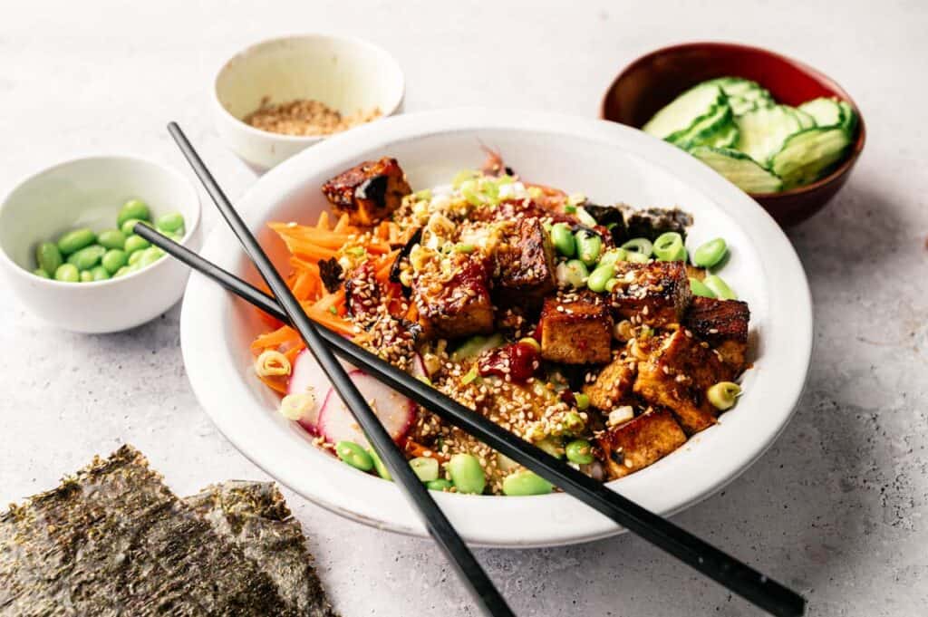 A vibrant Vegan Poke Bowl featuring tofu, edamame, rice, seaweed, cucumbers, carrots, and sesame seeds, served with chopsticks and flavorful side dishes.