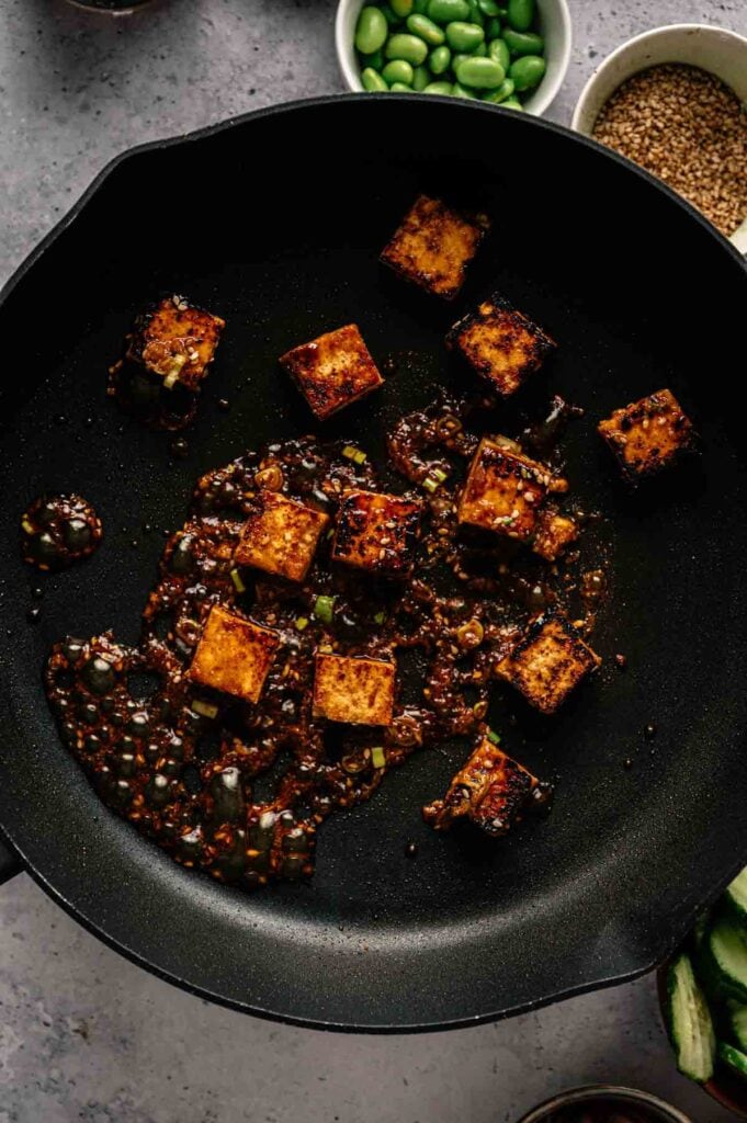 Cubes of tofu cooking in a black skillet with a dark, seasoned sauce&mdash;perfect for a Vegan Poke Bowl; small bowls of edamame and sesame seeds wait in the background.