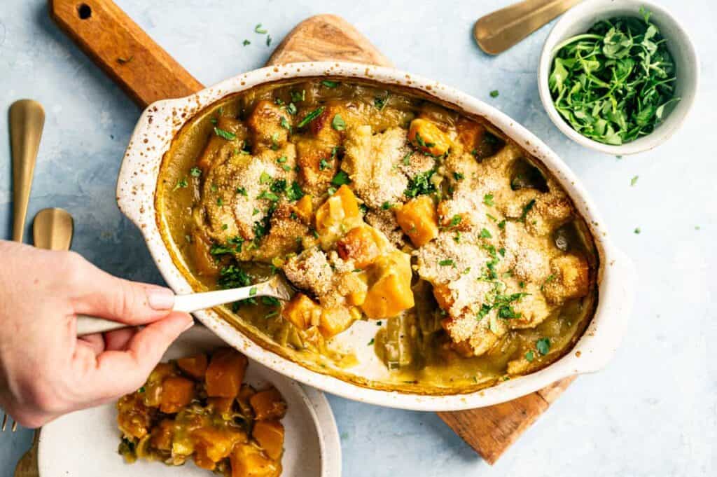 A hand serves a portion of Vegan Squash Casserole from a baked dish, topped with chopped herbs, onto a plate; nearby sits a bowl of fresh herbs.