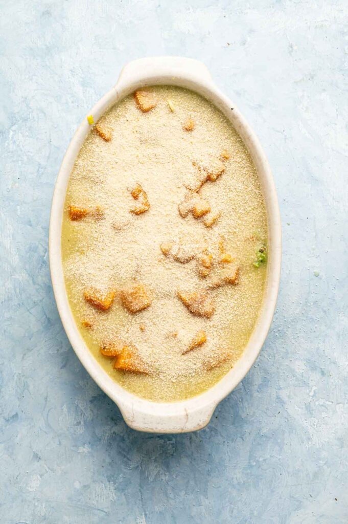 Oval baking dish filled with a creamy Vegan Squash Casserole, topped with bread crumbs and visible chunks of orange vegetable, on a light blue textured surface.