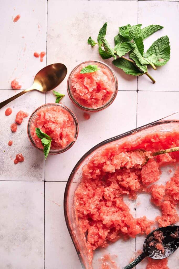 Glass dish and two jars of red watermelon sorbet granita garnished with mint leaves, a gold spoon, and mint sprigs on a white tiled surface.