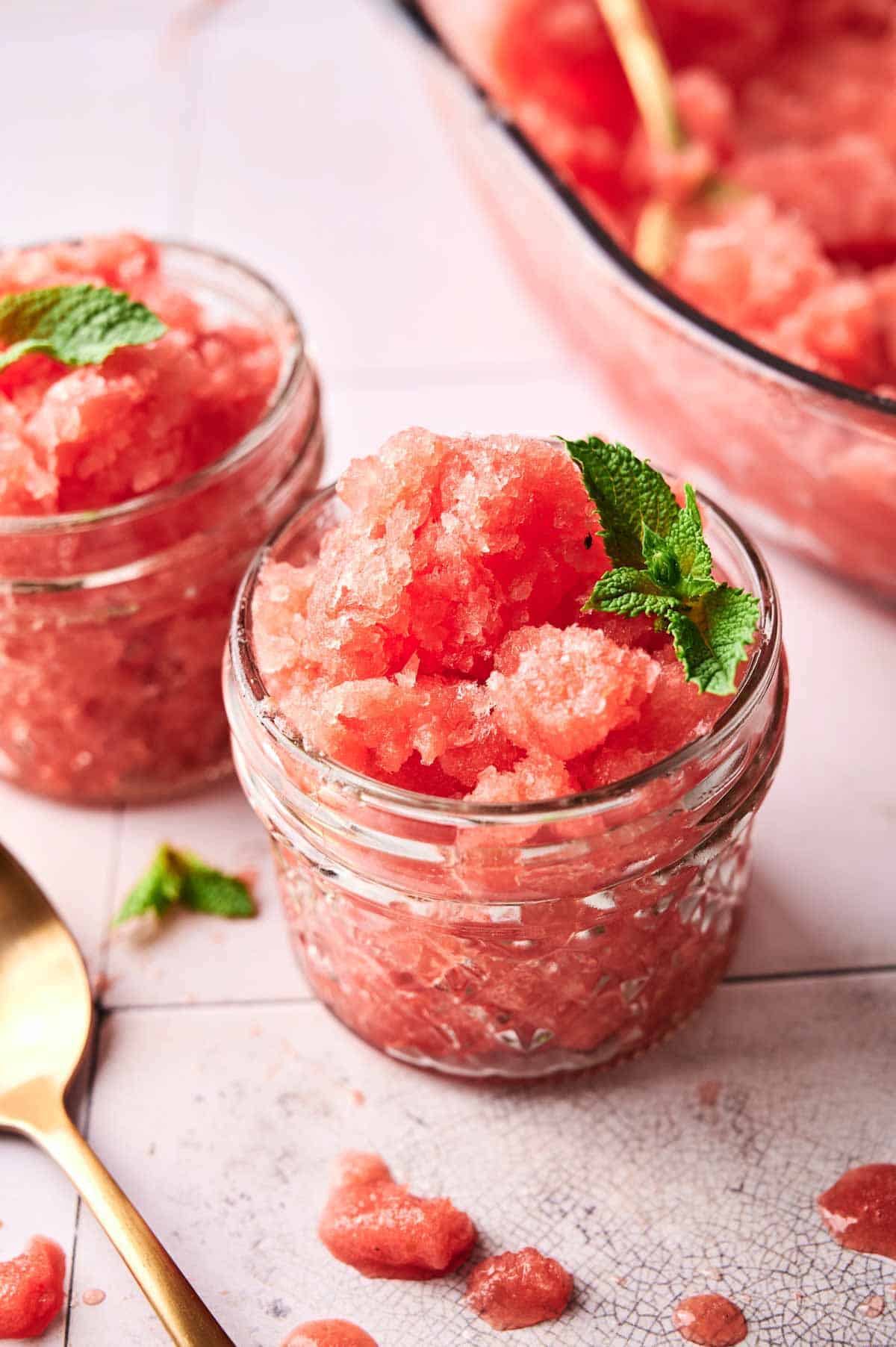 Two small glass jars filled with pink watermelon sorbet granita, garnished with fresh mint leaves. A spoon and some spilled sorbet are visible on a white tiled surface.