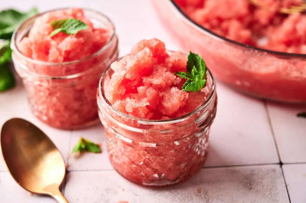 Two small glass jars filled with pink watermelon granita, reminiscent of refreshing Watermelon Sorbet, each garnished with a sprig of fresh mint, sit beside a gold spoon on a light-colored surface.