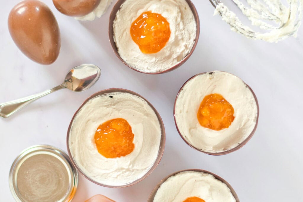 Four bowls of whipped cream topped with orange jam are arranged on a white surface, alongside a spoon, half chocolate egg, and a jar of jam&mdash;perfect as a light Vegan Easter Dessert for your holiday celebrations.