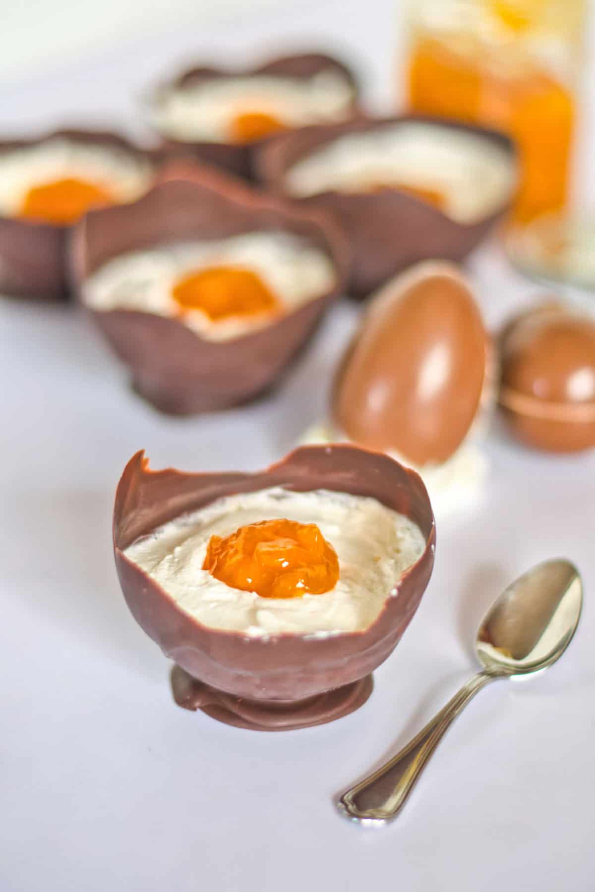Vegan Easter Dessert: Chocolate egg-shaped bowls filled with cream and topped with orange jam, with a spoon and whole chocolate eggs beside them on a white surface.