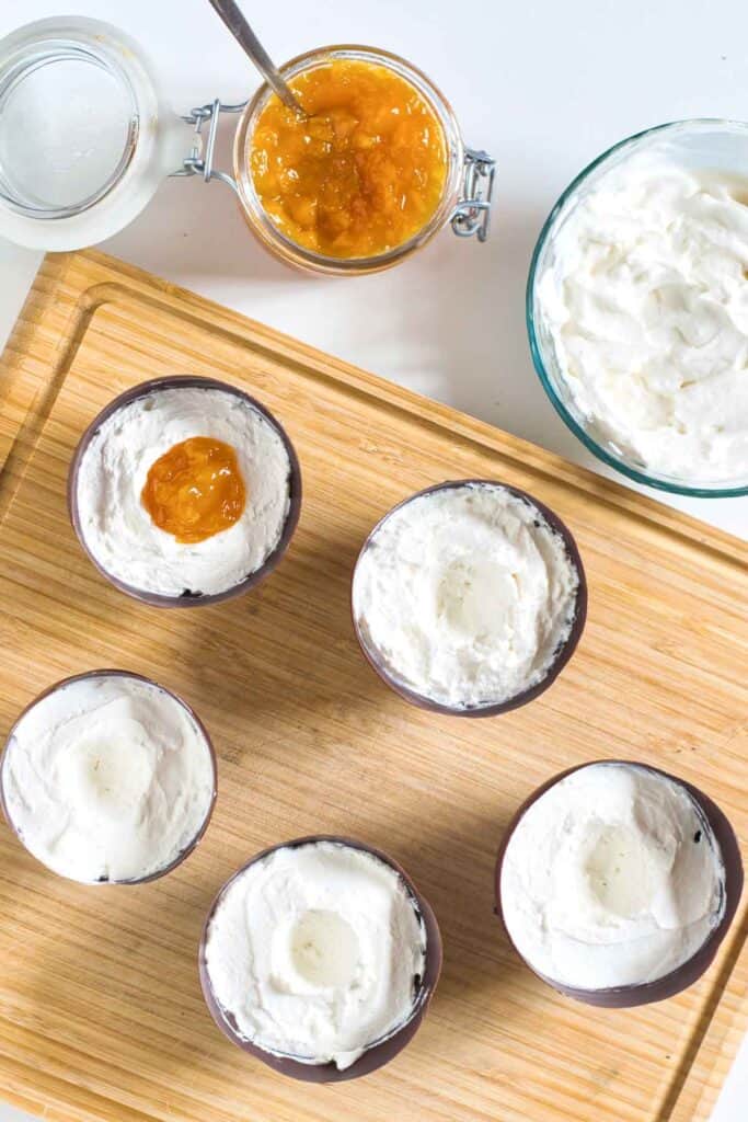 Six Vegan Easter Dessert cups with white cream, some with a cavity in the center&mdash;one filled with orange jam&mdash;are arranged on a wooden board next to a jar of jam and a bowl of whipped cream.