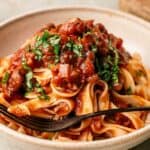 A plate of tagliatelle pasta topped with rich Mushroom Bolognese, fresh tomato sauce, chopped herbs, and a black fork resting on the side.