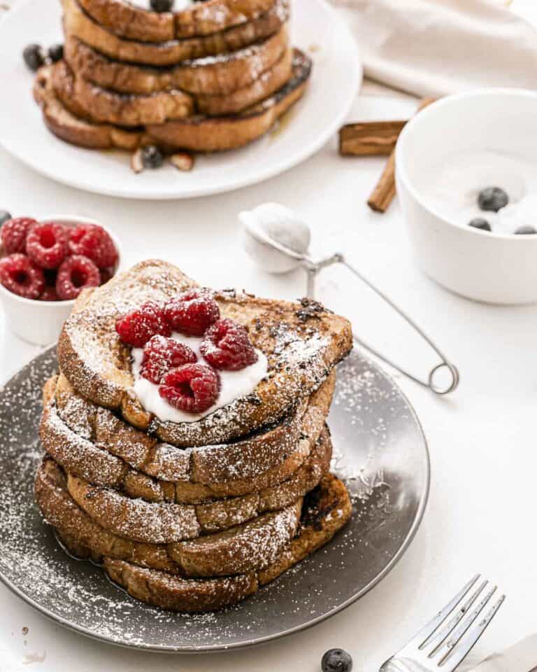 A stack of Vegan French Toast topped with whipped cream and raspberries sits on a gray plate, surrounded by fresh berries, a bowl of yogurt, and utensils on a white table.