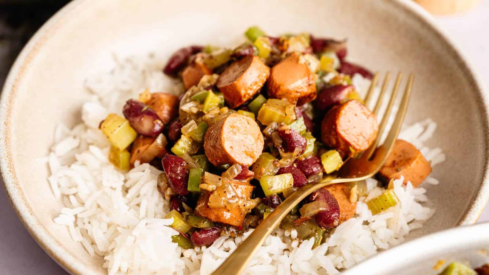 A bowl of white rice topped with sliced sausage, kidney beans, chopped celery, and onions, with a gold fork resting on the side.