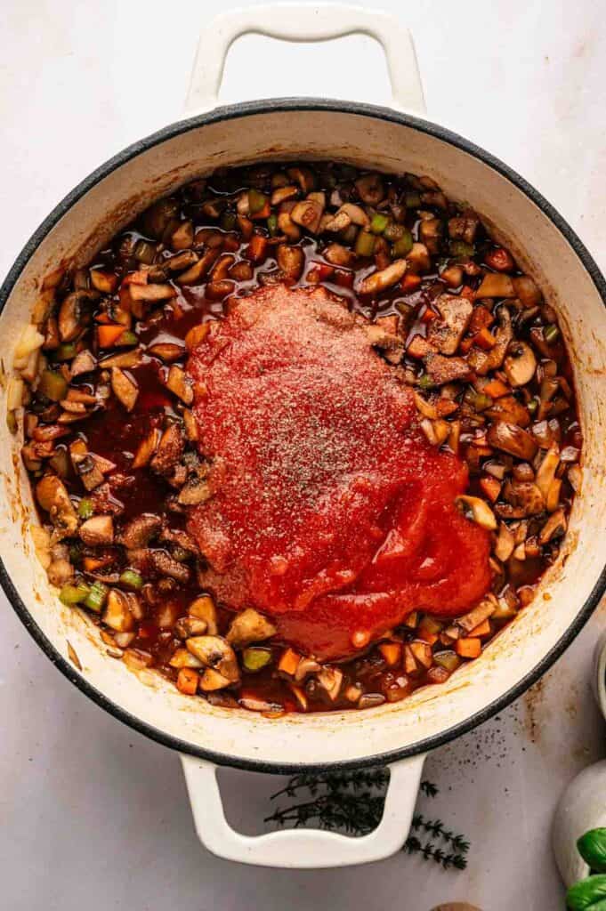 A white pot filled with chopped vegetables, tomato sauce, and seasonings for a flavorful Mushroom Bolognese sits on a white surface, ready to be stirred and cooked.