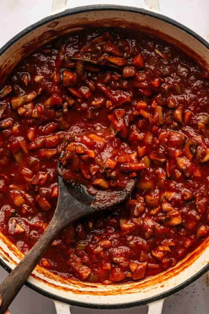 A pot of chunky Mushroom Bolognese, with a rich tomato-based sauce and visible pieces of vegetables, being stirred with a wooden spoon.