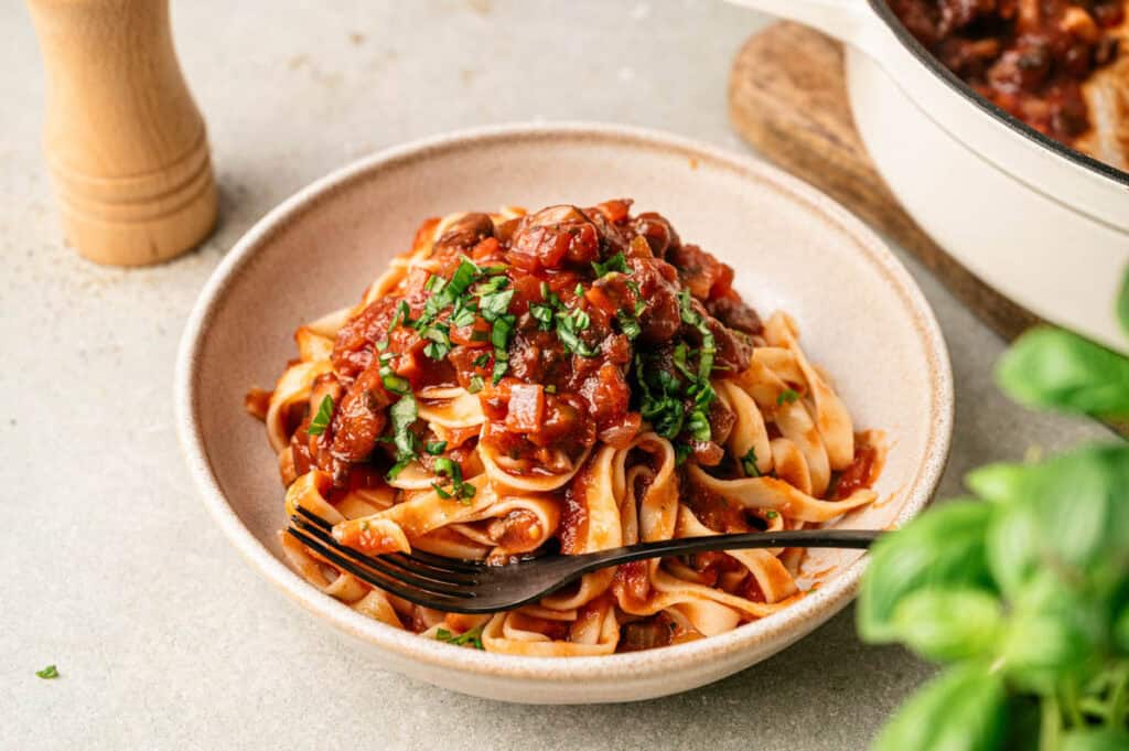 A bowl of tagliatelle pasta topped with rich Mushroom Bolognese sauce and chopped herbs, with a fork resting on the side. A pepper grinder and a pot are visible in the background.