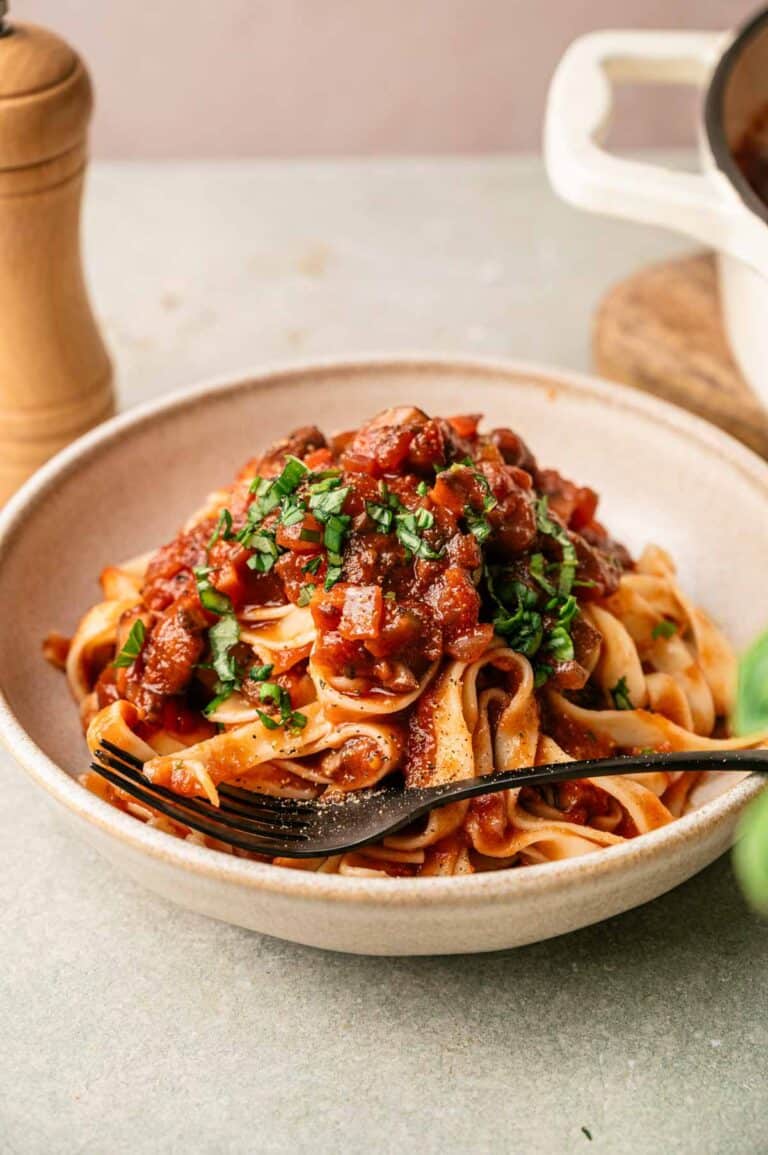 A bowl of tagliatelle pasta topped with rich Mushroom Bolognese sauce and chopped fresh herbs, with a fork resting on the side.