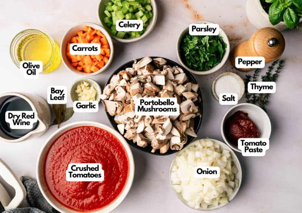 A variety of labeled ingredients for a Mushroom Bolognese are arranged on a countertop, including chopped portobello mushrooms, onion, celery, carrots, parsley, garlic, and bowls of crushed tomatoes and seasonings.