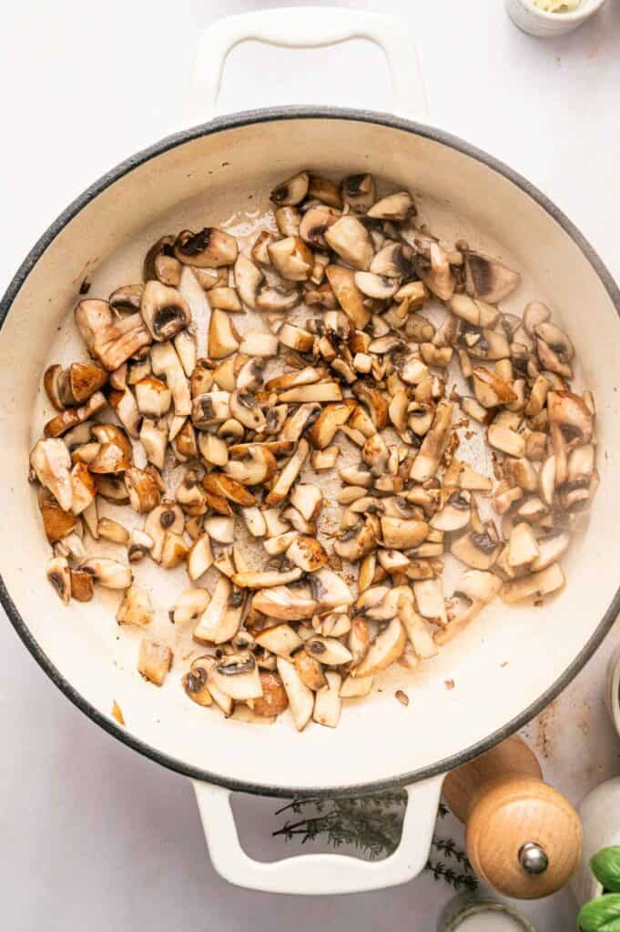 Chopped mushrooms being sautéed in a large white pan for a savory Mushroom Bolognese, with some steam rising, surrounded by herbs and seasoning on a white surface.