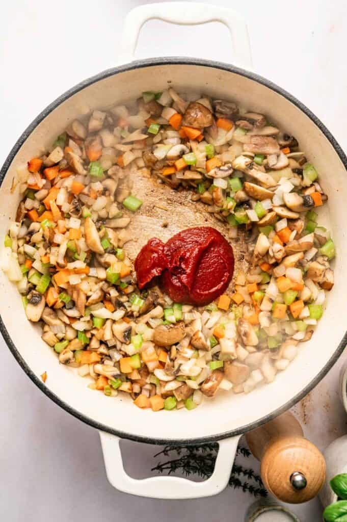 Diced celery, carrots, onions, and mushrooms sauté in a white pot with a dollop of tomato paste in the center—perfect for starting a hearty Mushroom Bolognese.